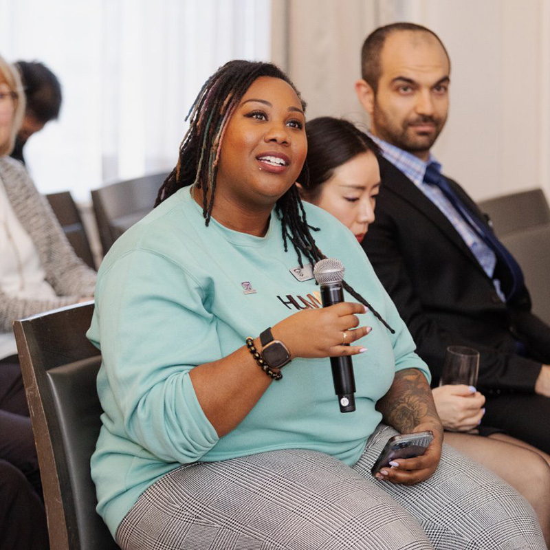 Person seated at a conference holding a microphone and a smartphone, with other attendees sitting nearby..